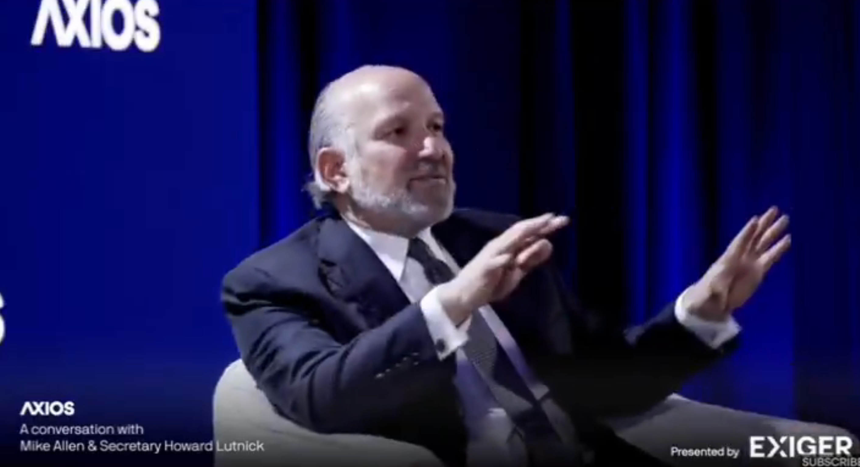 A man in a business suit gestures while sitting on stage at an Axios event, engaging in a discussion