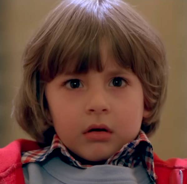 A young child with wide eyes, wearing a plaid shirt under a jacket, looking surprised or alarmed