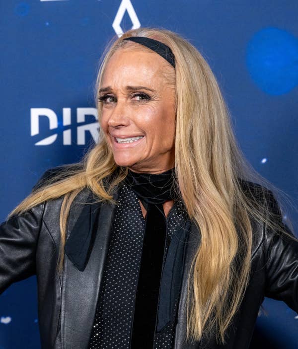 Person smiling at an event, wearing a leather jacket and scarf, with long hair held back by a headband
