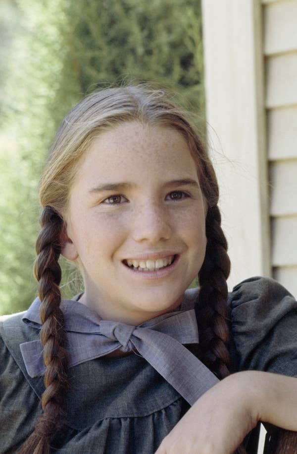 Person with braided hair smiling, wearing vintage-style dress with a large bow, outdoors