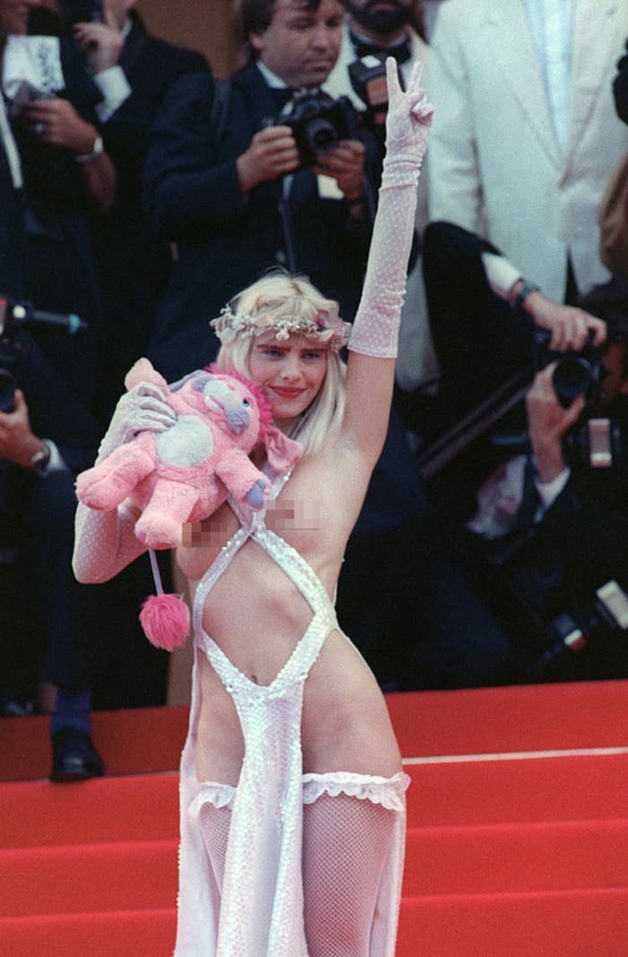 La Cicciolina on a red carpet in an avant-garde outfit that's just a cross cross of fabric exposing her breasts, holding a plush toy, posing with one arm raised, surrounded by photographers