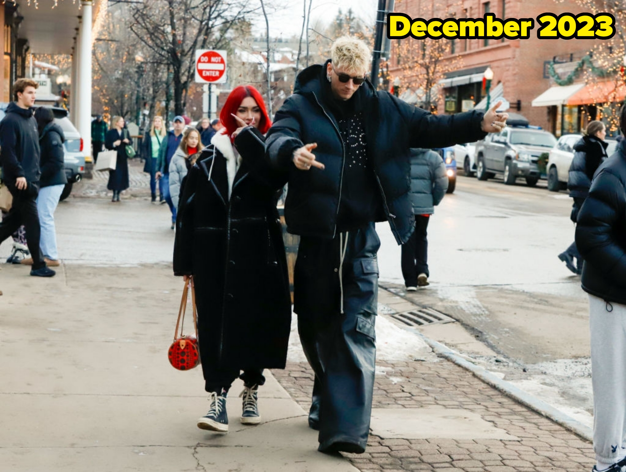 Two people walking on a city street in winter, wearing stylish, oversized black coats and sunglasses. Trees with lights in the background