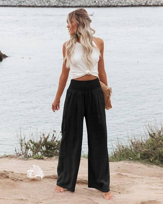 model successful  sleeveless apical  and wide-leg pants lasting  barefoot by the water, holding a clutch, showcasing a casual chic outfit