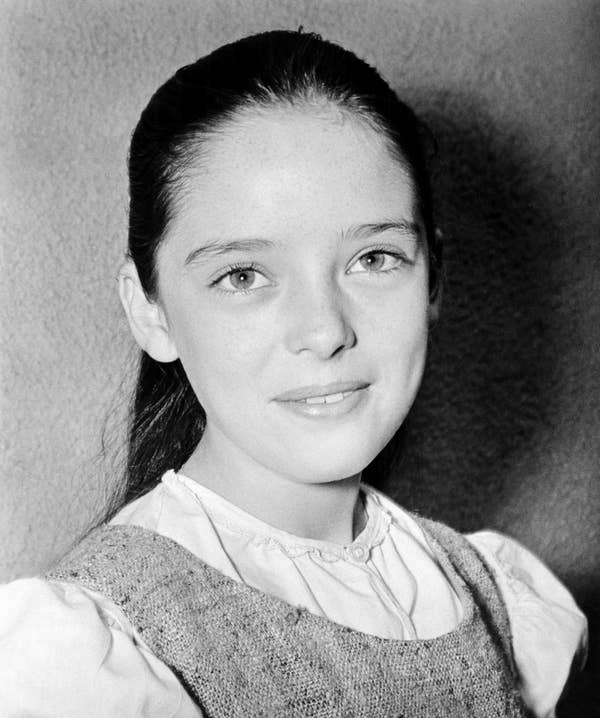 Young girl with long hair and a gentle smile, wearing a light blouse under a textured dress, looking at the camera.