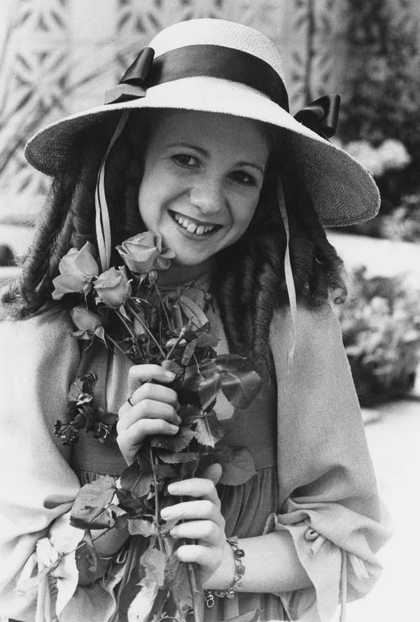 Person in vintage attire with a wide-brimmed hat holds a bouquet of roses, smiling warmly