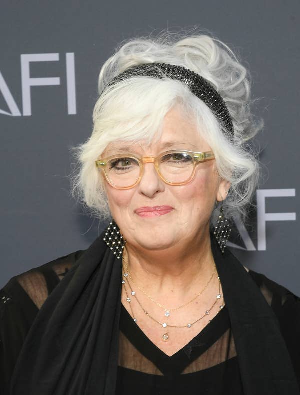 A person with white hair styled up, wearing glasses, a black headband, and a sheer black top with layered necklaces at a celebrity event