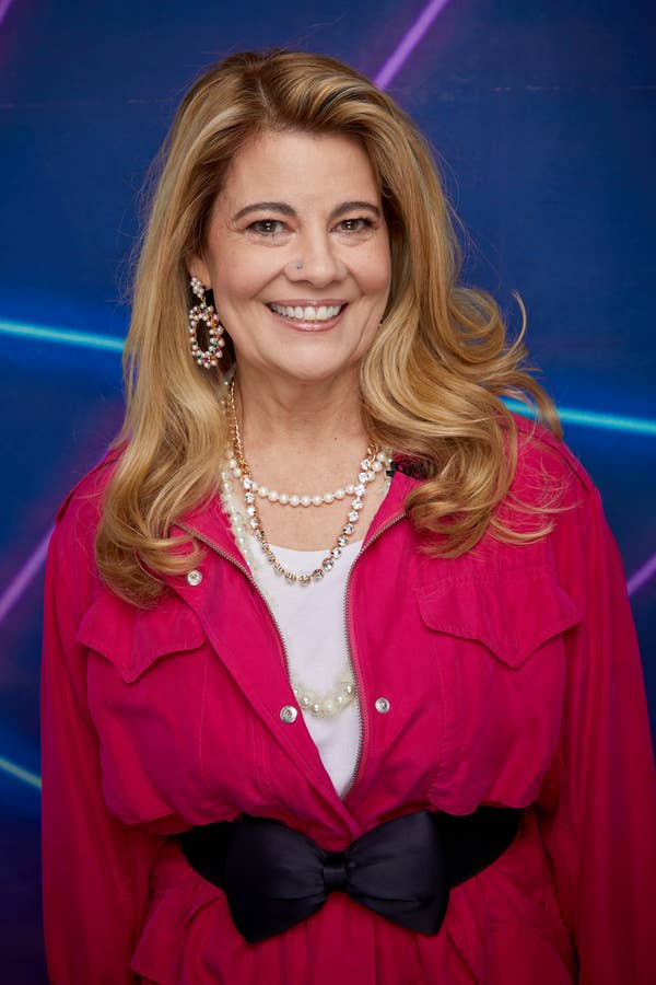 A woman with long hair wearing large hoop earrings, pearl necklaces, a white top, a vibrant jacket, and a wide belt stands against a neon backdrop