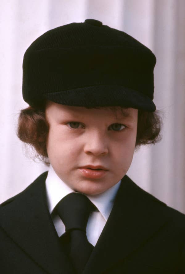 A child in formal attire with a black cap and tie stands facing the camera