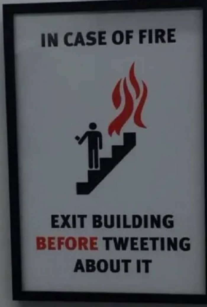 Sign jokingly advises to exit the building before tweeting in case of fire, featuring a stick figure near flames on stairs
