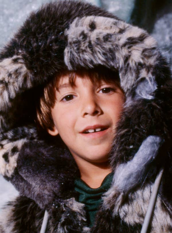 A person dressed in a fur-lined hooded coat stands against an icy backdrop, smiling