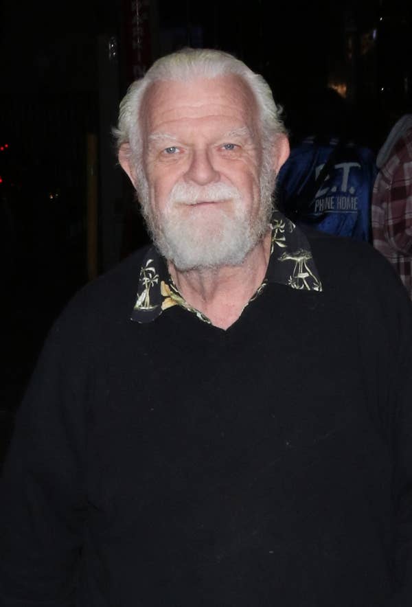 An older man with white hair and beard walks down a city sidewalk at night, wearing a casual dark sweater and slacks