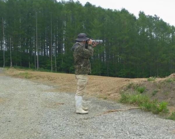 A person in outdoor gear, including a wide-brim hat and boots, takes a photo in a rural landscape with tall trees in the background