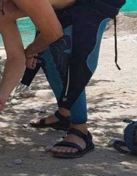 Person wearing a wetsuit and sandals stands on a sandy beach, adjusting their gear