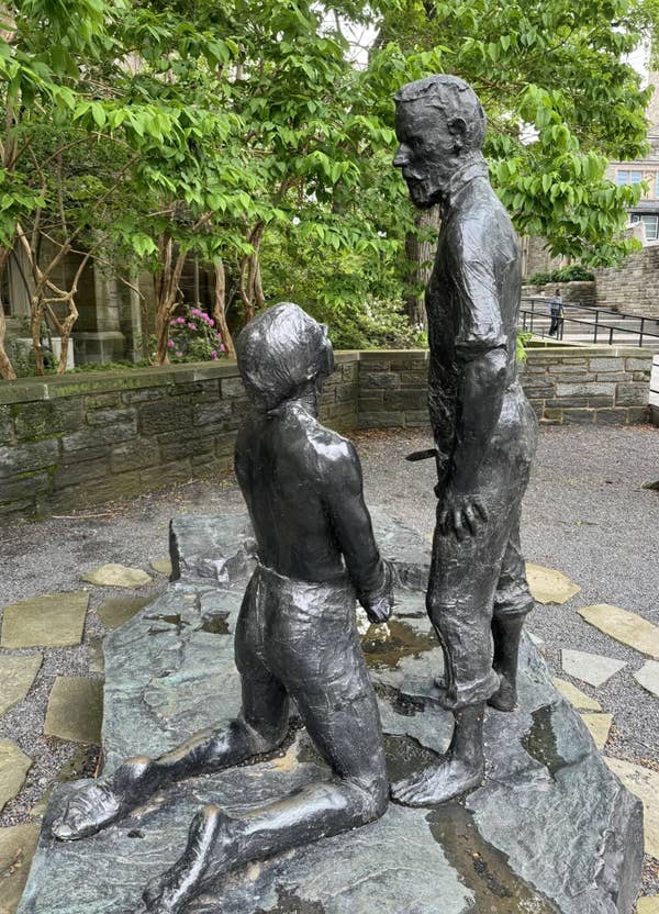 Bronze statue of a standing figure interacting with a figure kneeling on a stone base in an outdoor setting with greenery