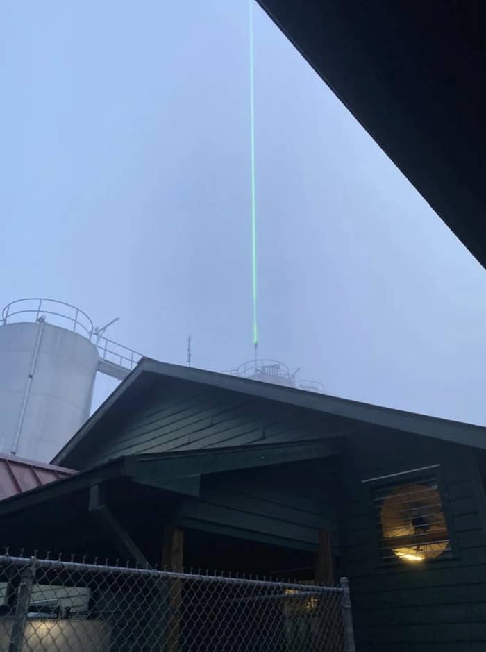 A green laser beam shoots upward from rooftops against a foggy sky, with industrial tanks visible in the background