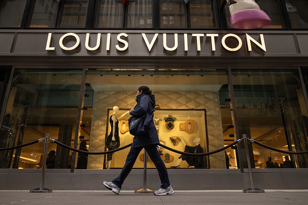 A person walks past a Louis Vuitton store with fashion displays in the window, roped off with stanchions