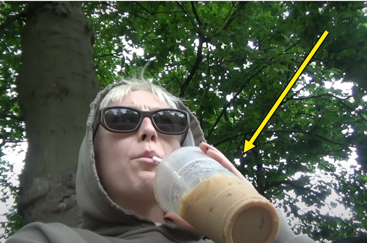 Person with short hair and sunglasses in a hoodie drinks iced coffee through a straw under trees. Arrow points to the drink