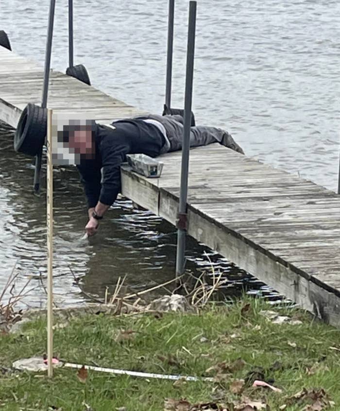 Person in a hoodie and pants lies on a dock reaching into the water, near grassy shore and reeds