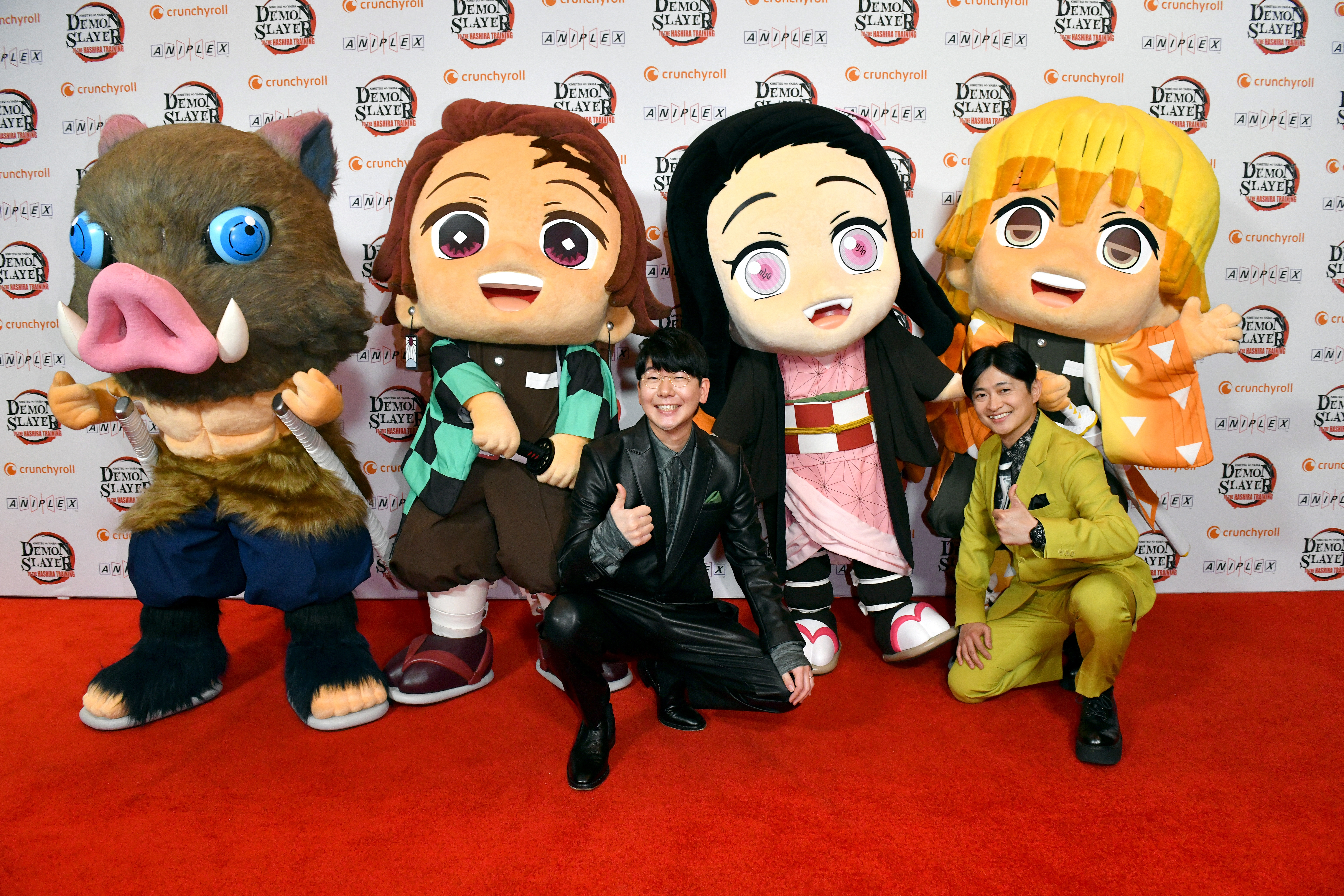 People pose with large anime character mascots in playful costumes on a red carpet, two individuals give thumbs up and smile at the camera
