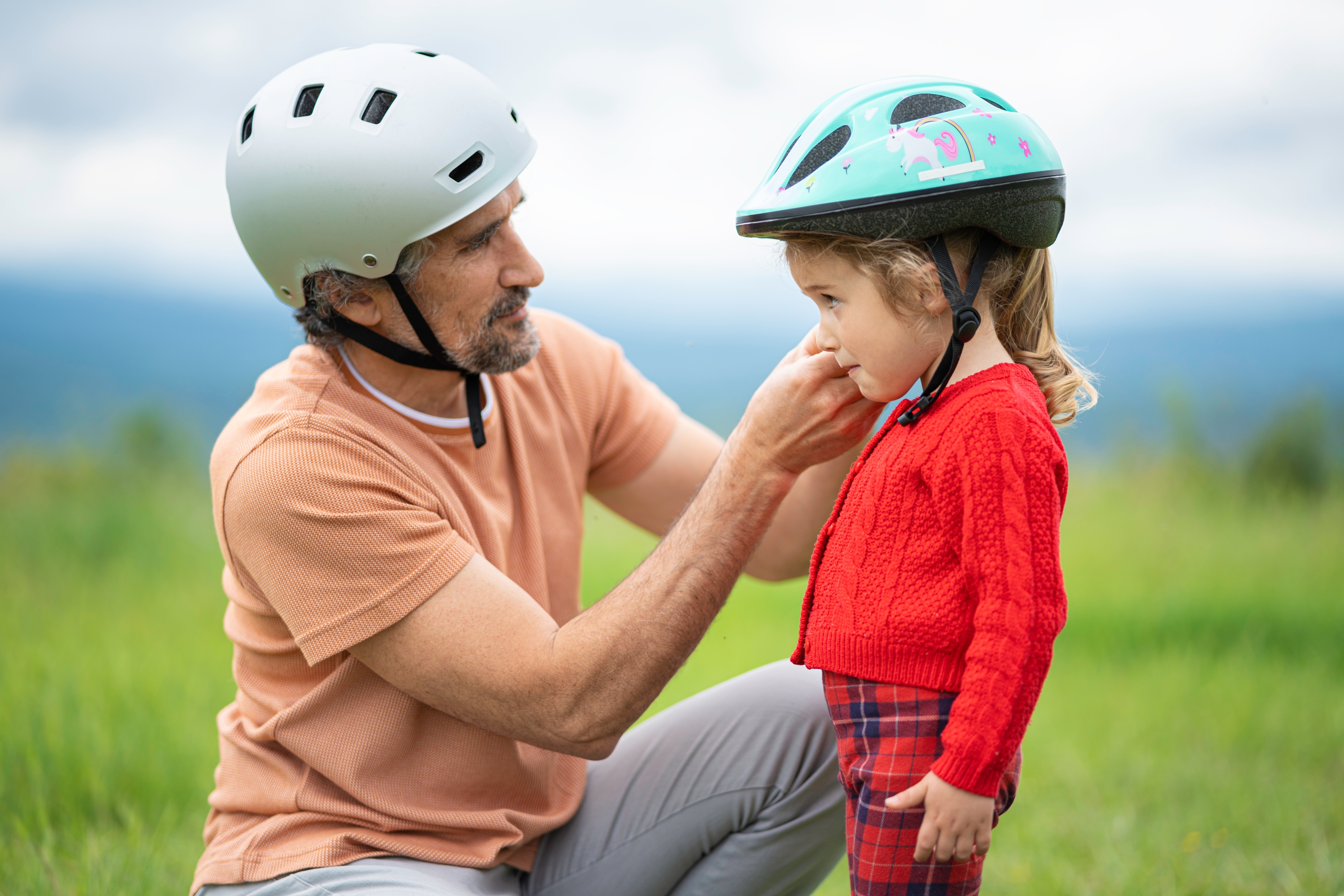 An adult helps a child fasten a bicycle helmet in an outdoor setting, emphasizing safety and care