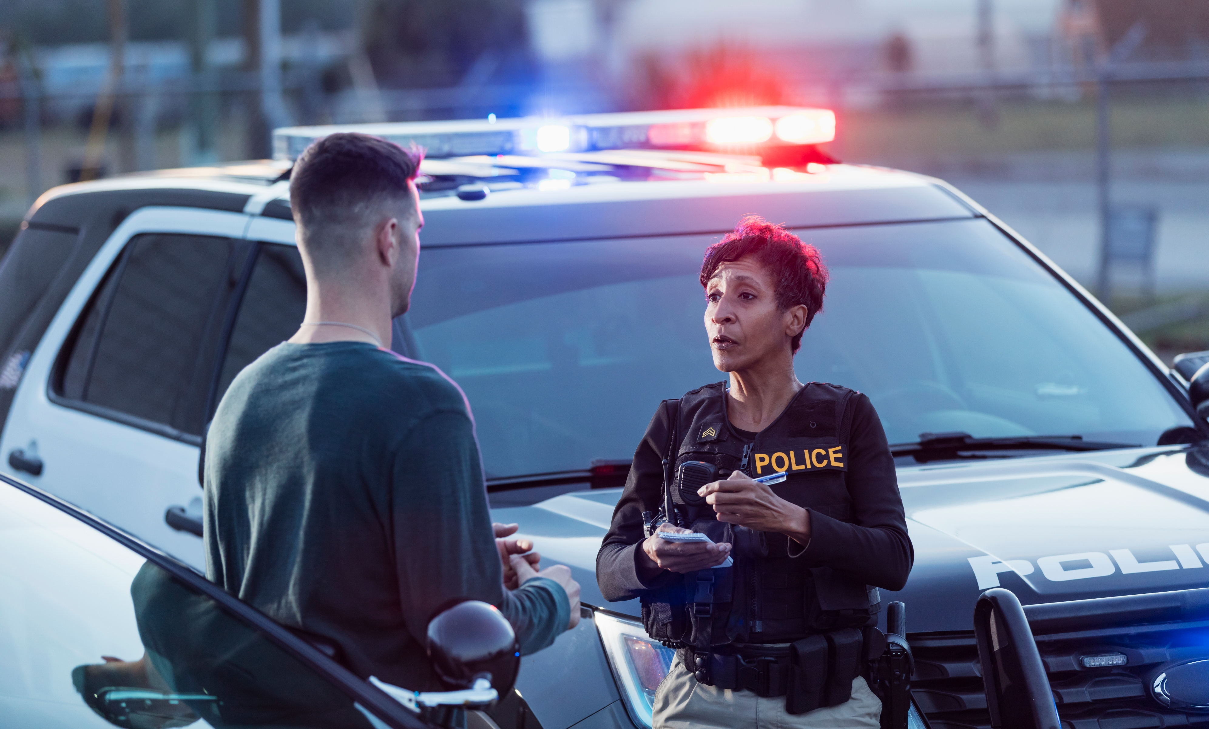 Police officer talks to a man beside a patrol car with flashing lights in an urban setting