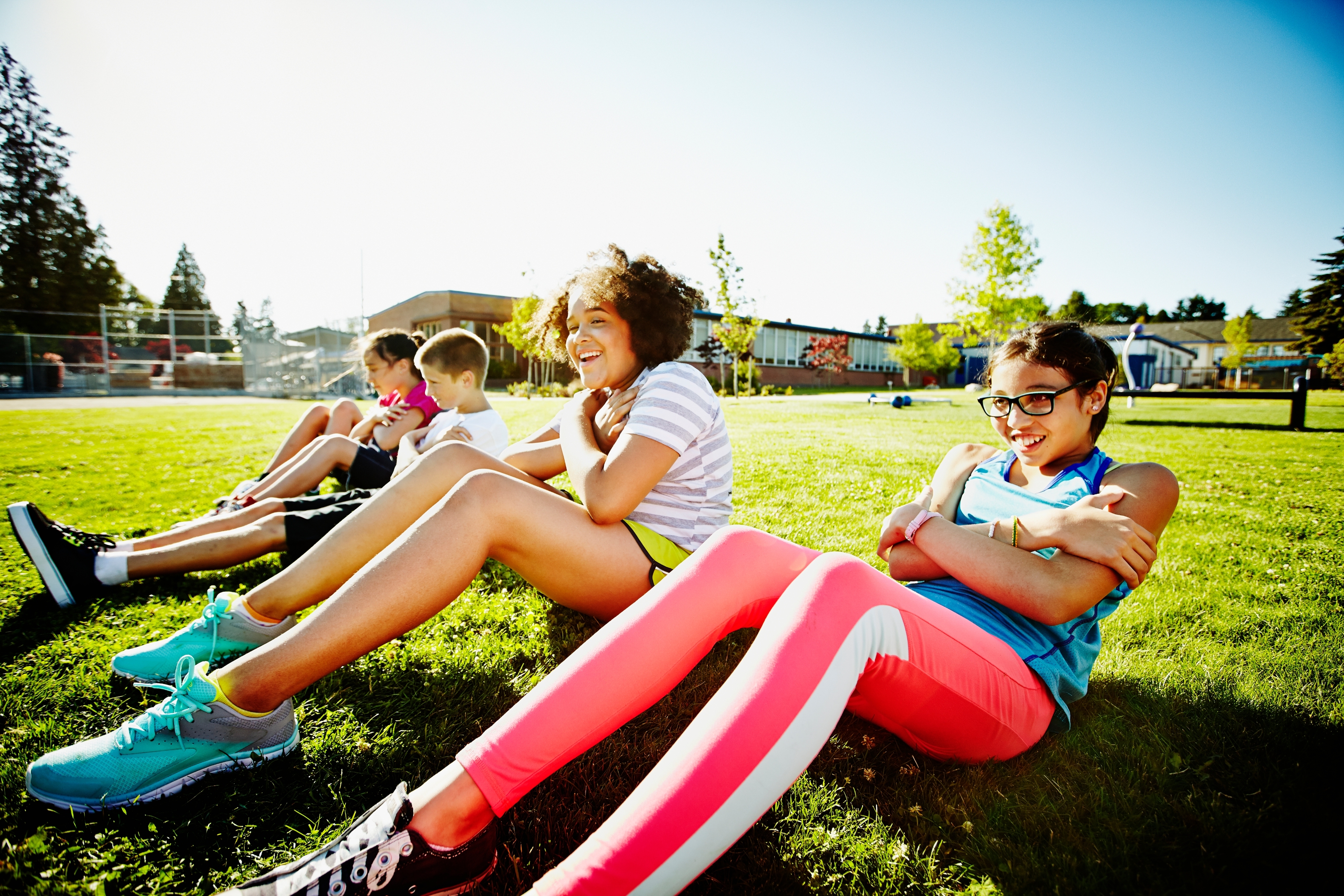 Group of kids sitting on grass in a park, laughing and doing sit-ups together during a sunny day
