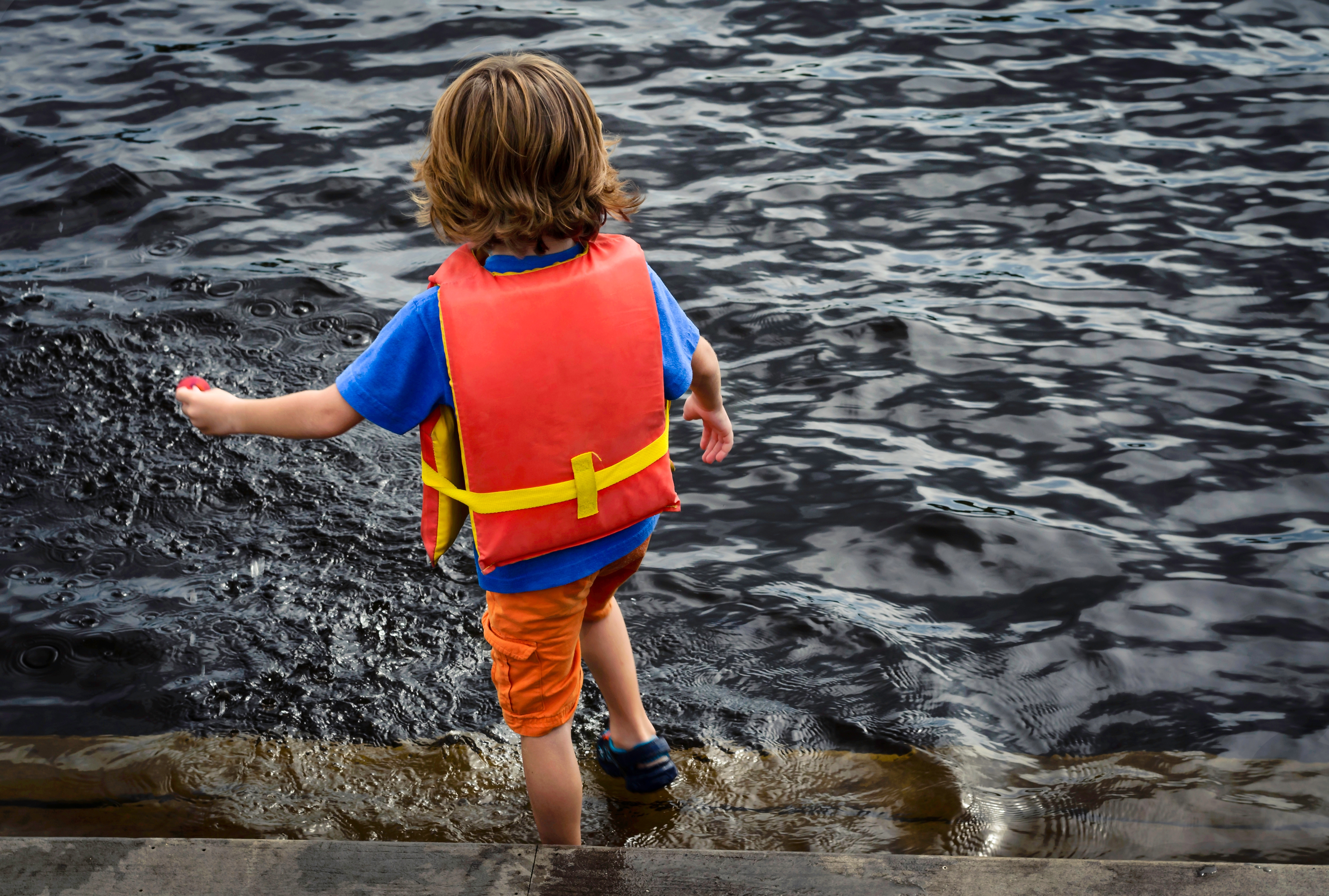 Barn i en redningsvest står ved vannets kant på en brygge, ser inn i vannet, iført uformelle sommerklær