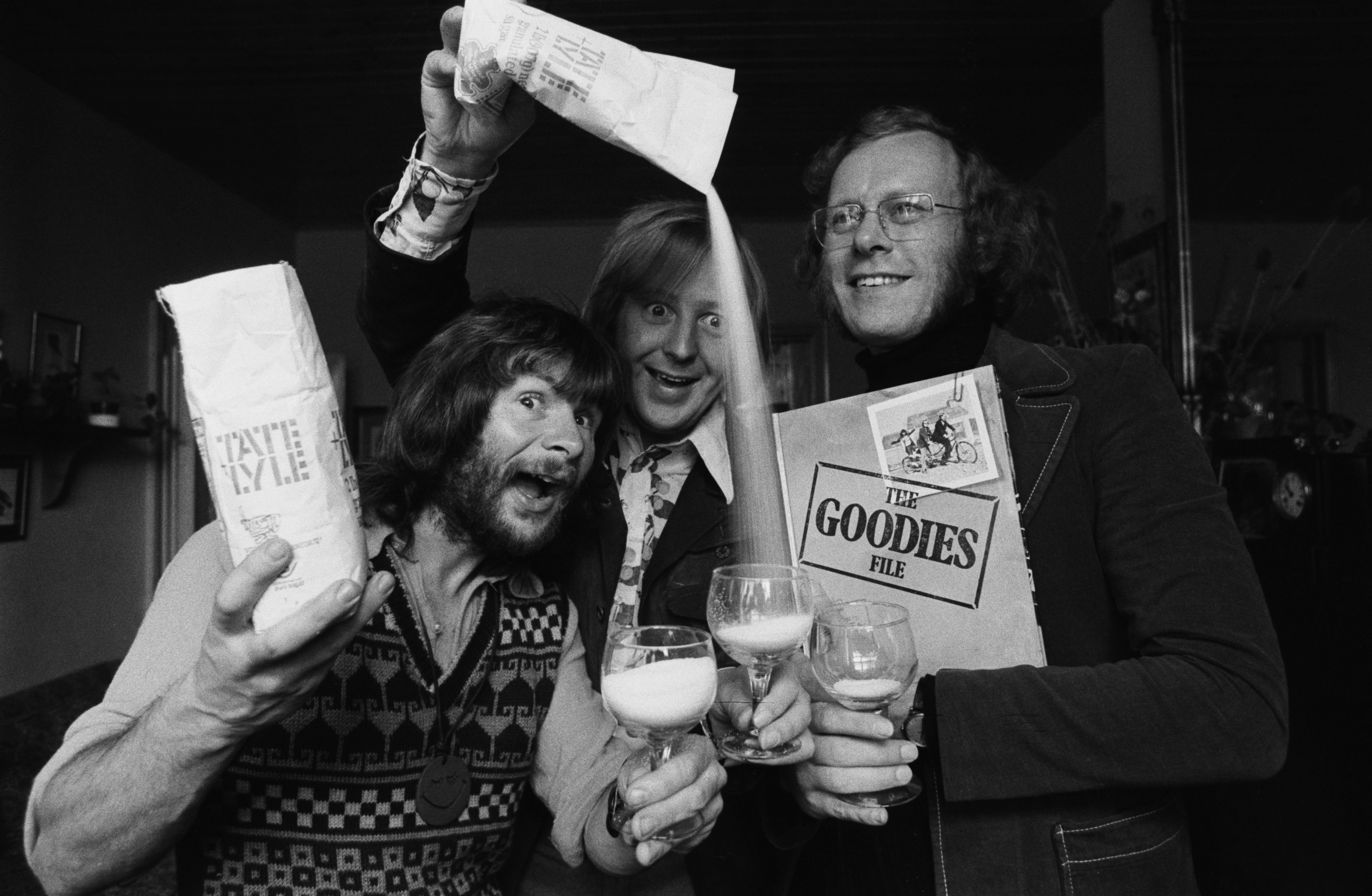 Three people joyfully pour cereal into glasses, each holding a box labeled "Tasty K-E-ye" and "The Goodies File," laughing in a playful manner