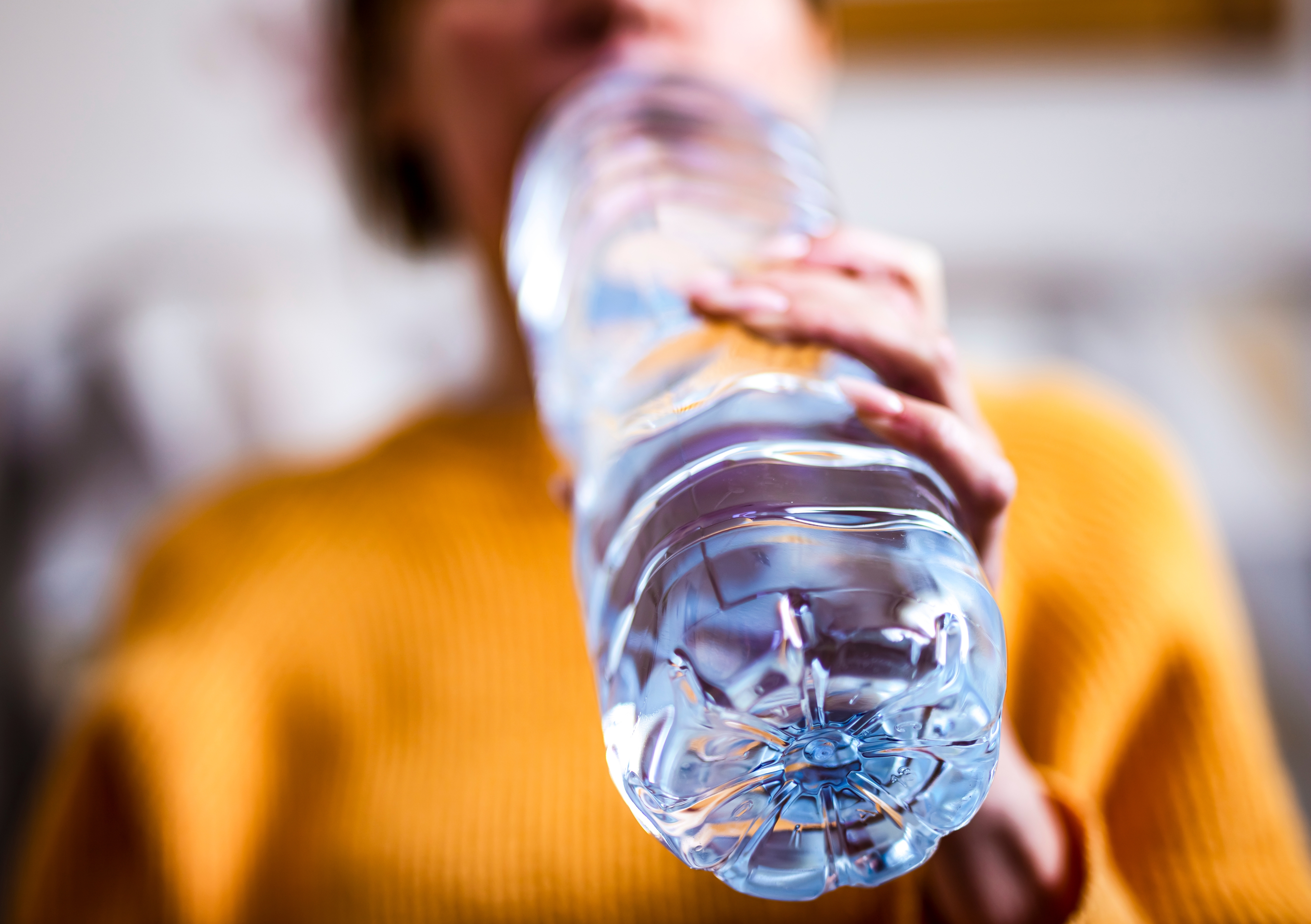 Person in a textured sweater drinks from a large, clear plastic bottle. The focus is on the bottle and hand