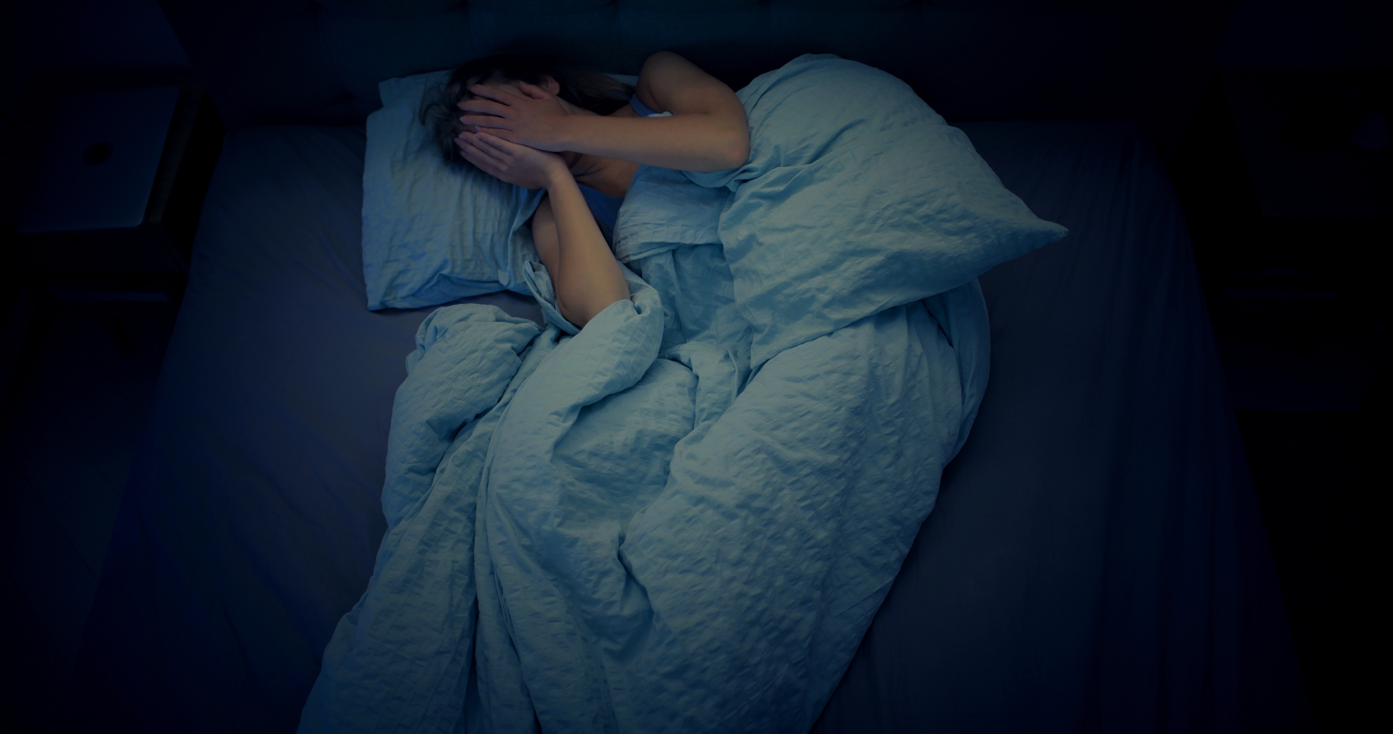 Person lying in bed with hands covering face, appearing stressed or upset, under crumpled bedding