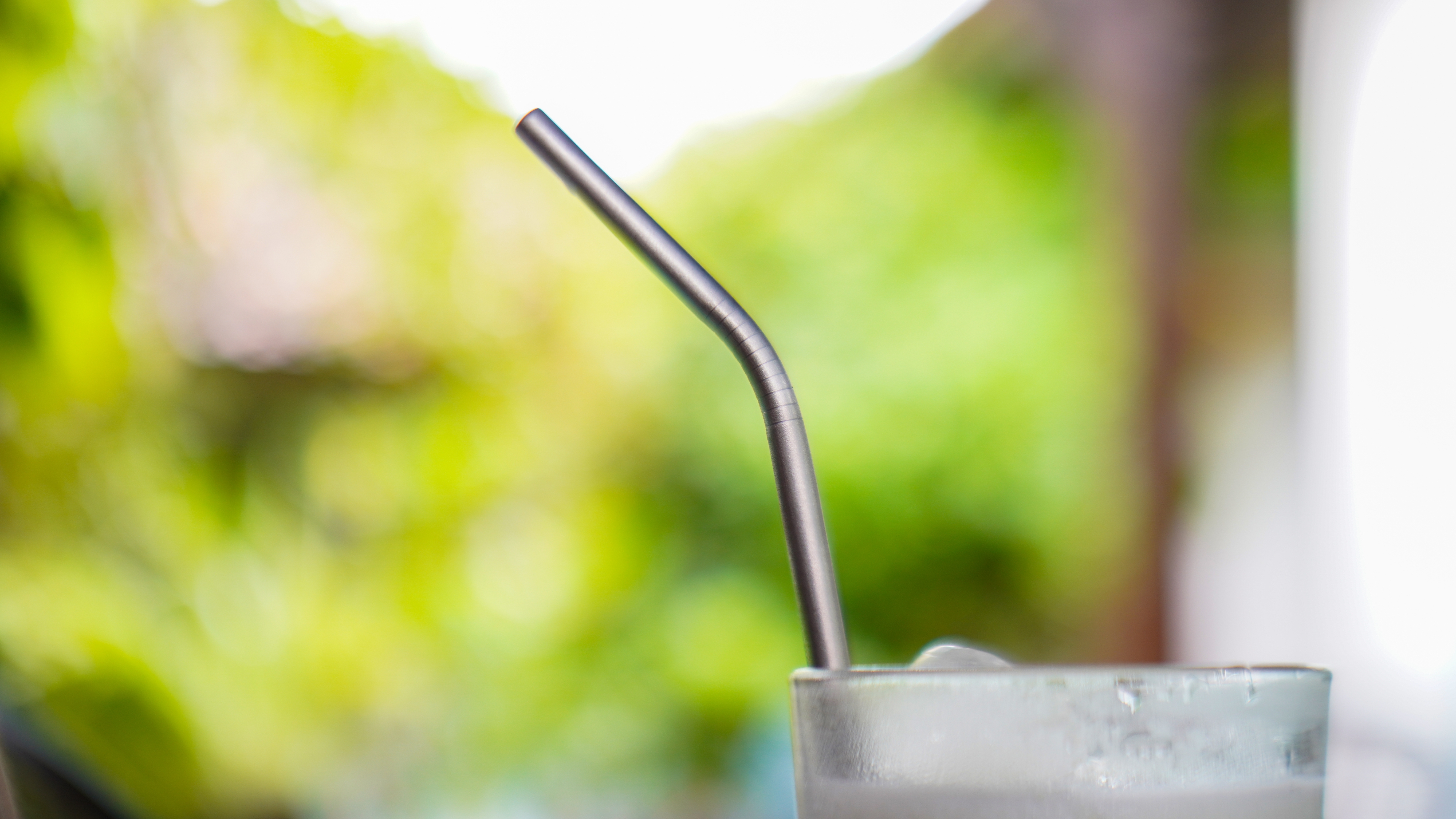 A metal straw is placed inside a frosted glass filled with a cold beverage, with a lush, blurred outdoor background