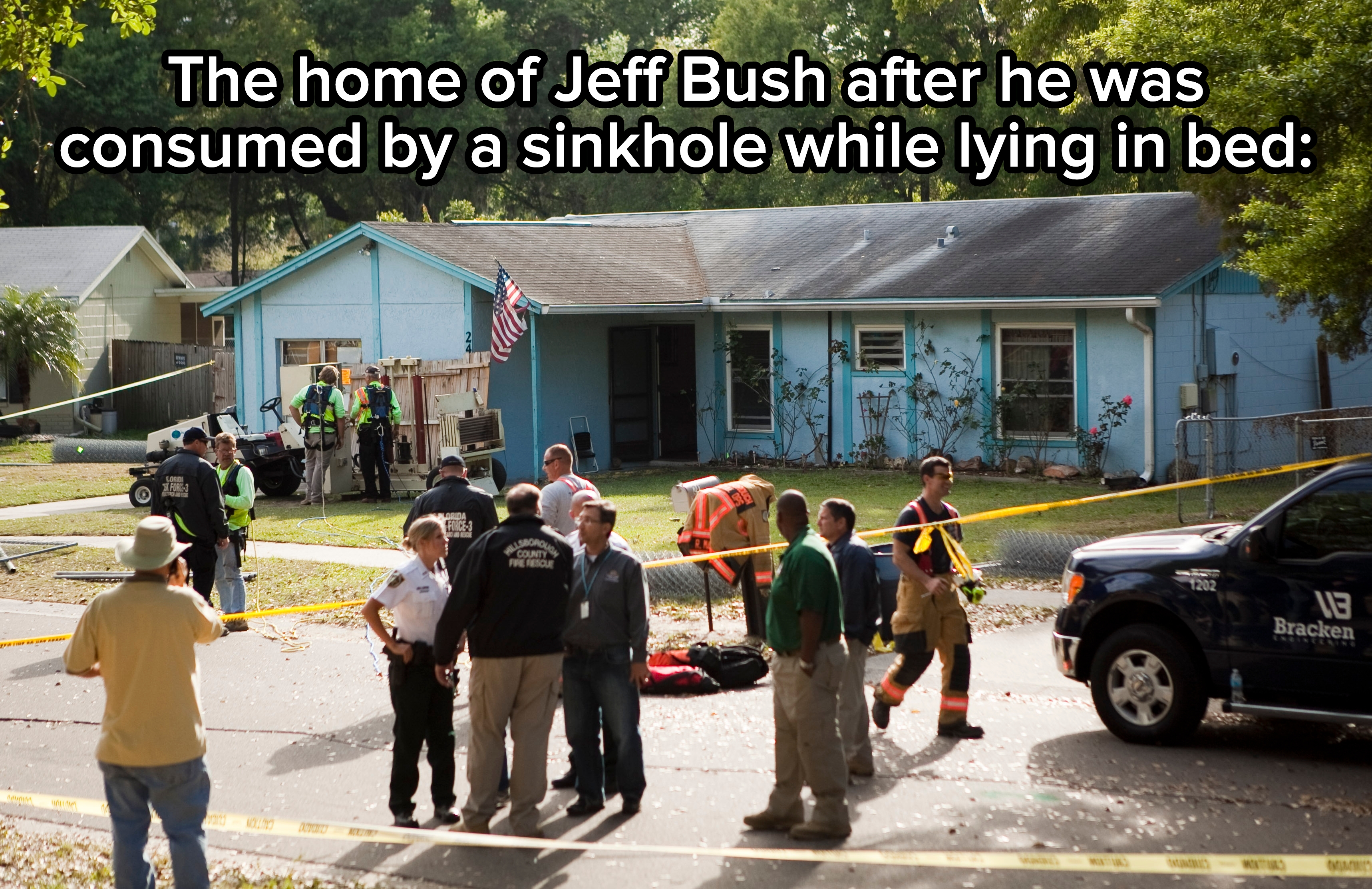 People gather outside a house with construction and police presence, surrounded by caution tape and a flag
