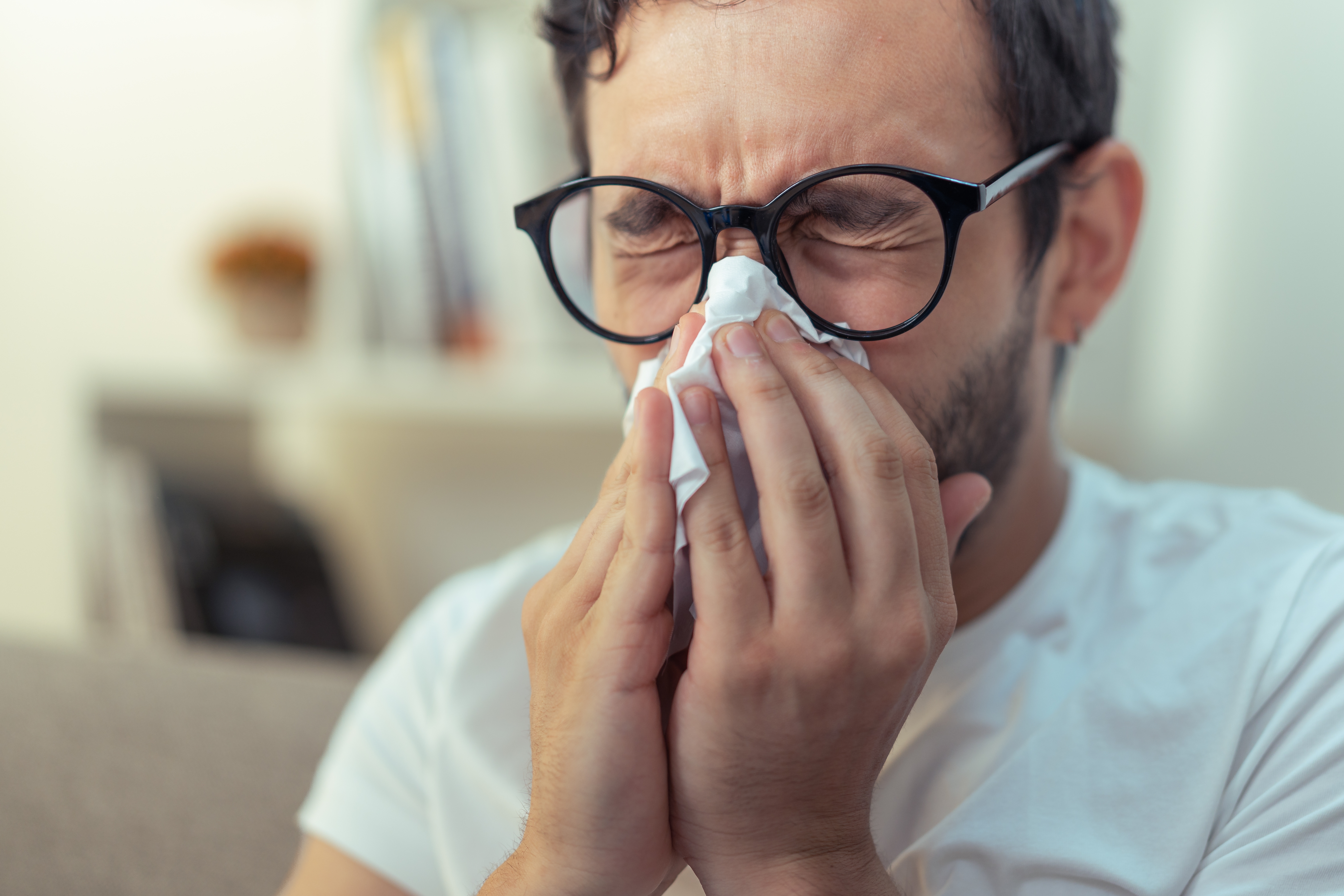 Person sneezing into a tissue with eyes closed, wearing glasses and a casual shirt