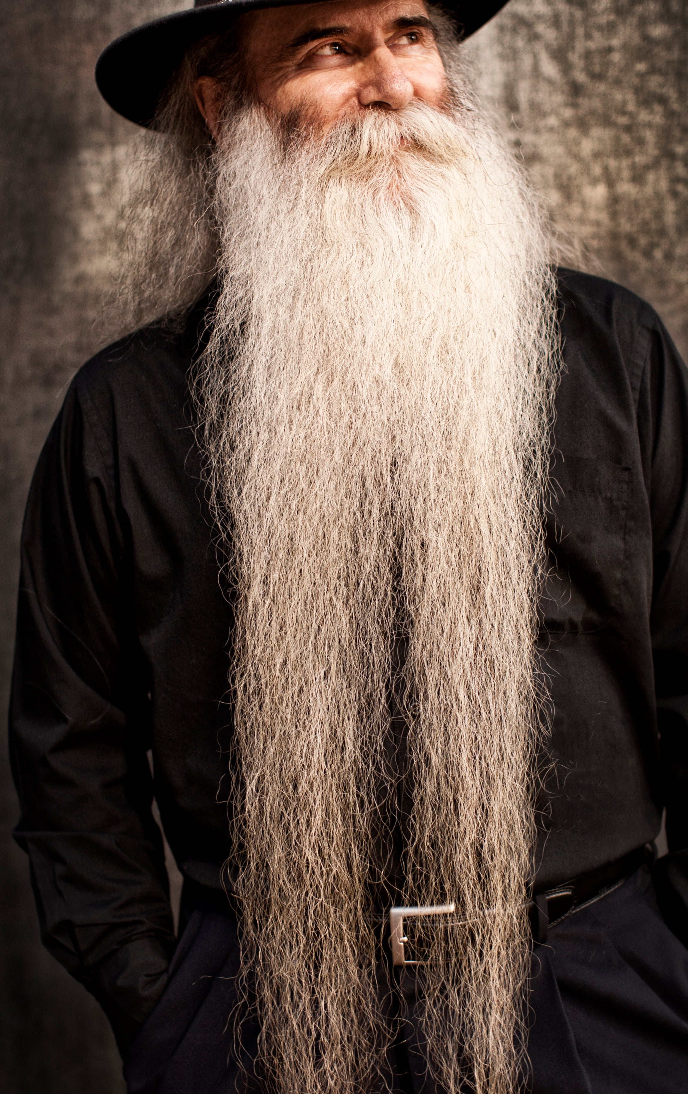 Man with a long white beard and mustache wears a black wide-brimmed hat and matching black outfit, smiling with hands in pockets
