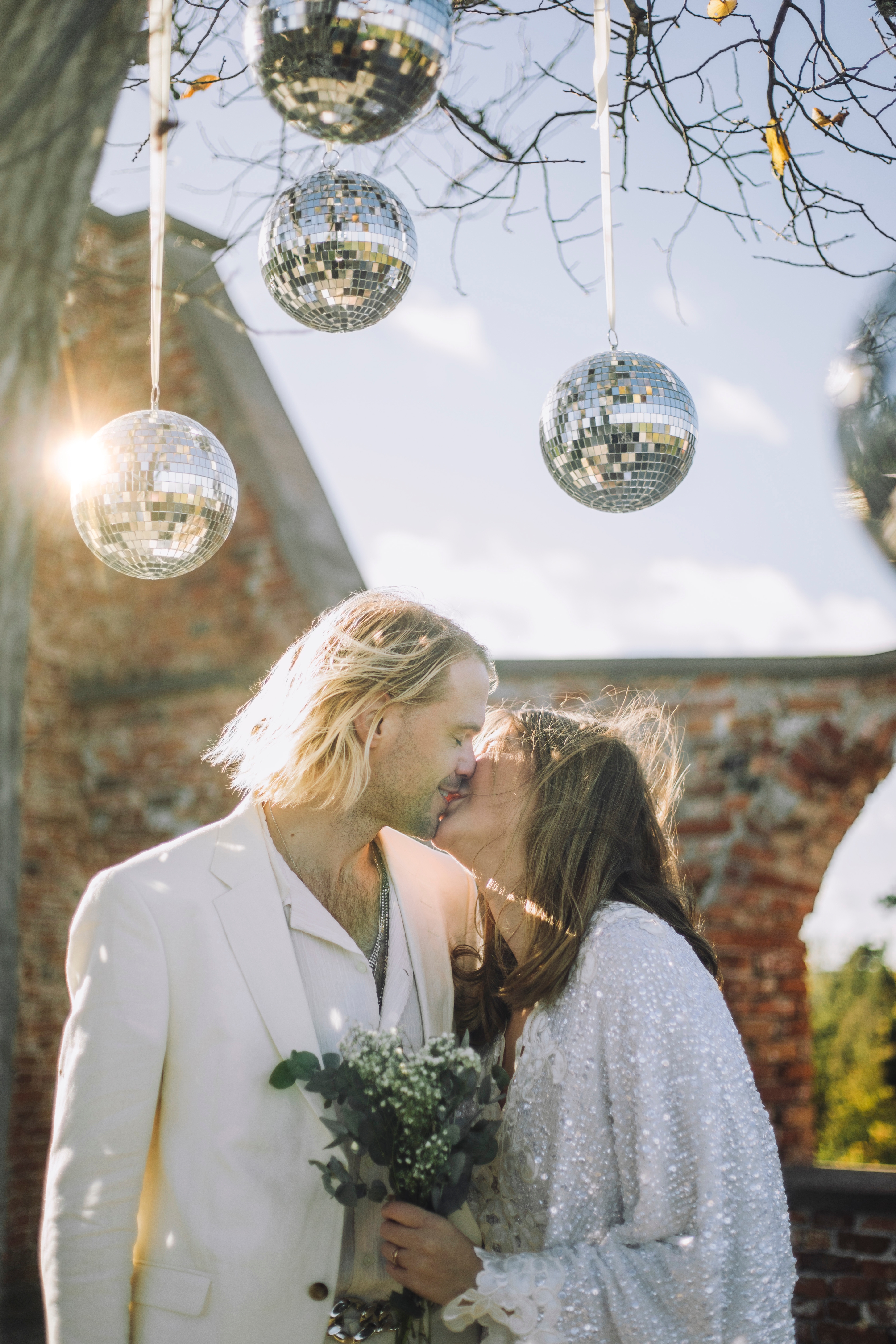 A couple kisses under disco balls; they wear stylish suits and hold a bouquet, creating a charming wedding ambiance