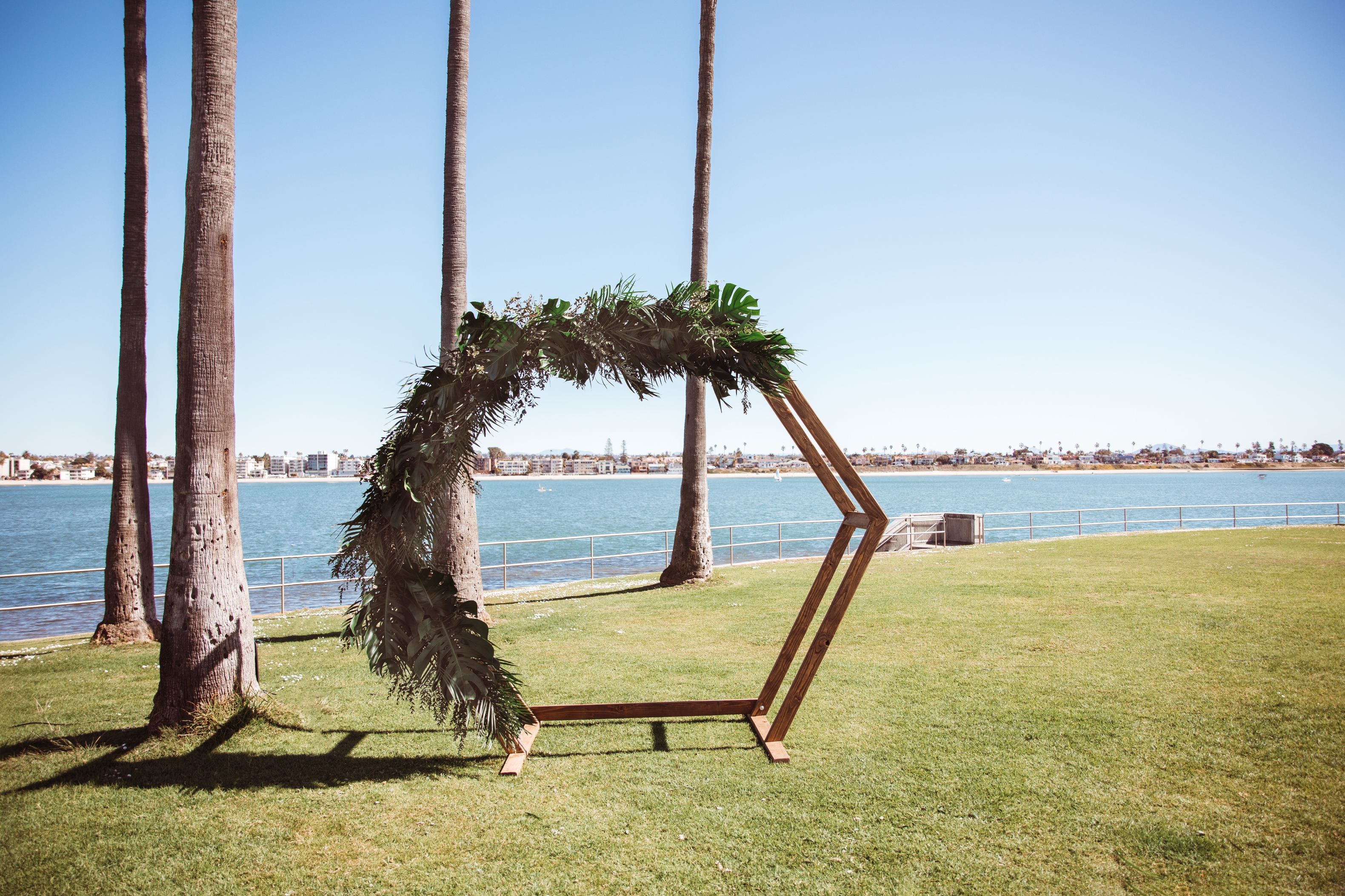 Hexagonal wooden wedding arch with greenery, set against a lakeside backdrop with palm trees and clear sky