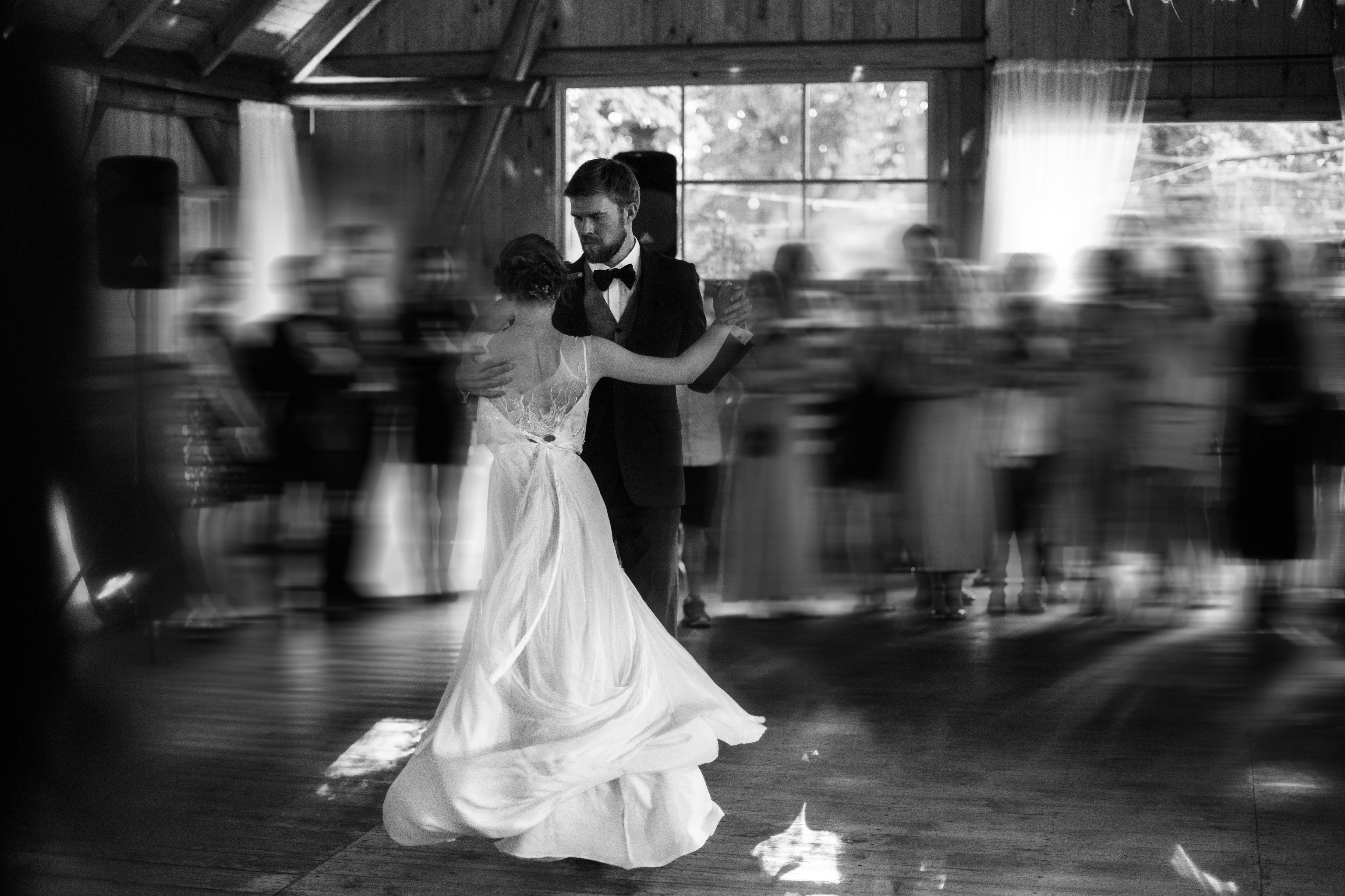 A couple performs a graceful dance at their wedding reception in a rustic venue, surrounded by blurred, cheering guests