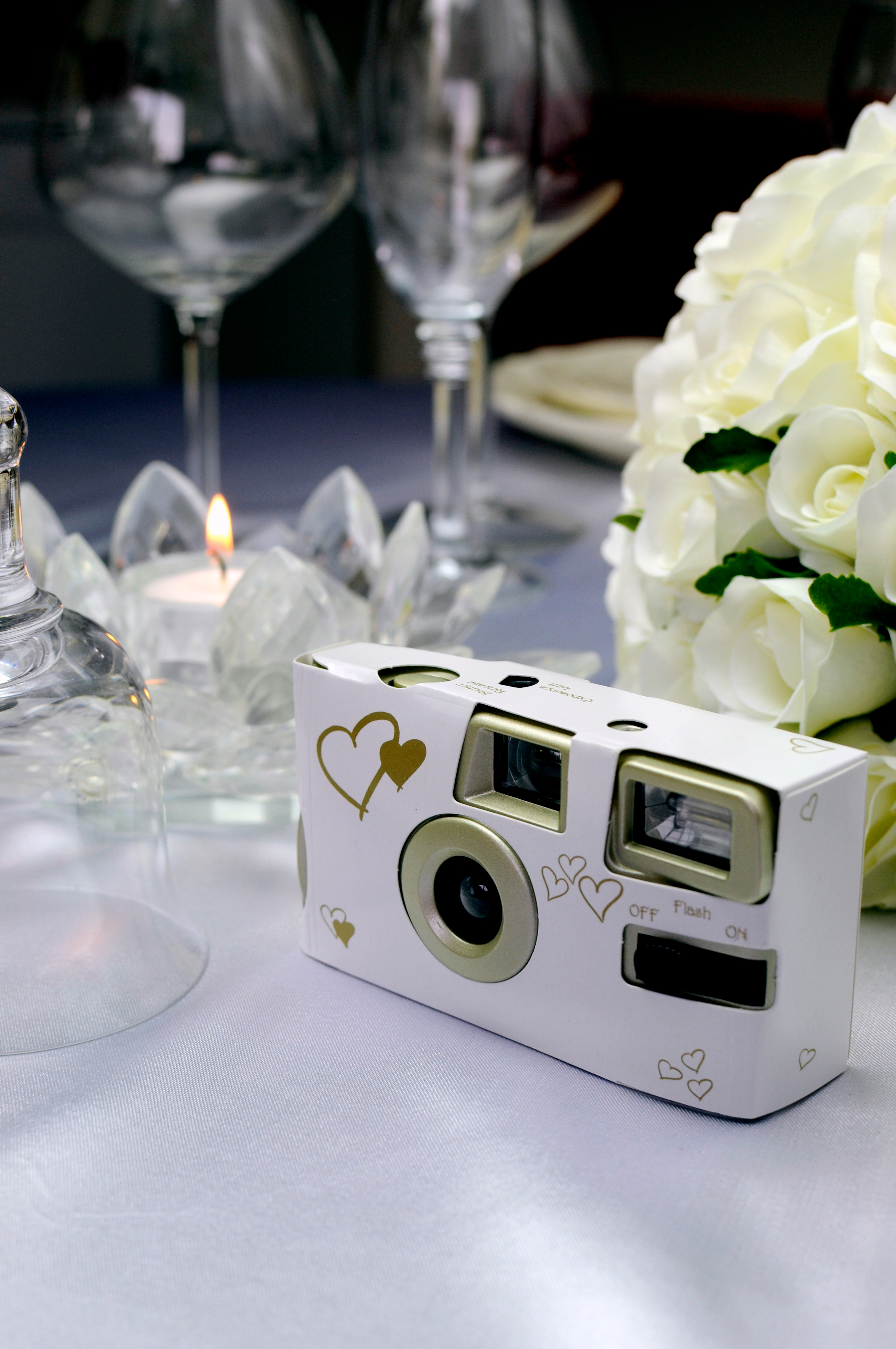Disposable camera with heart designs on a wedding reception table, surrounded by wine glasses, a candle, and white flowers