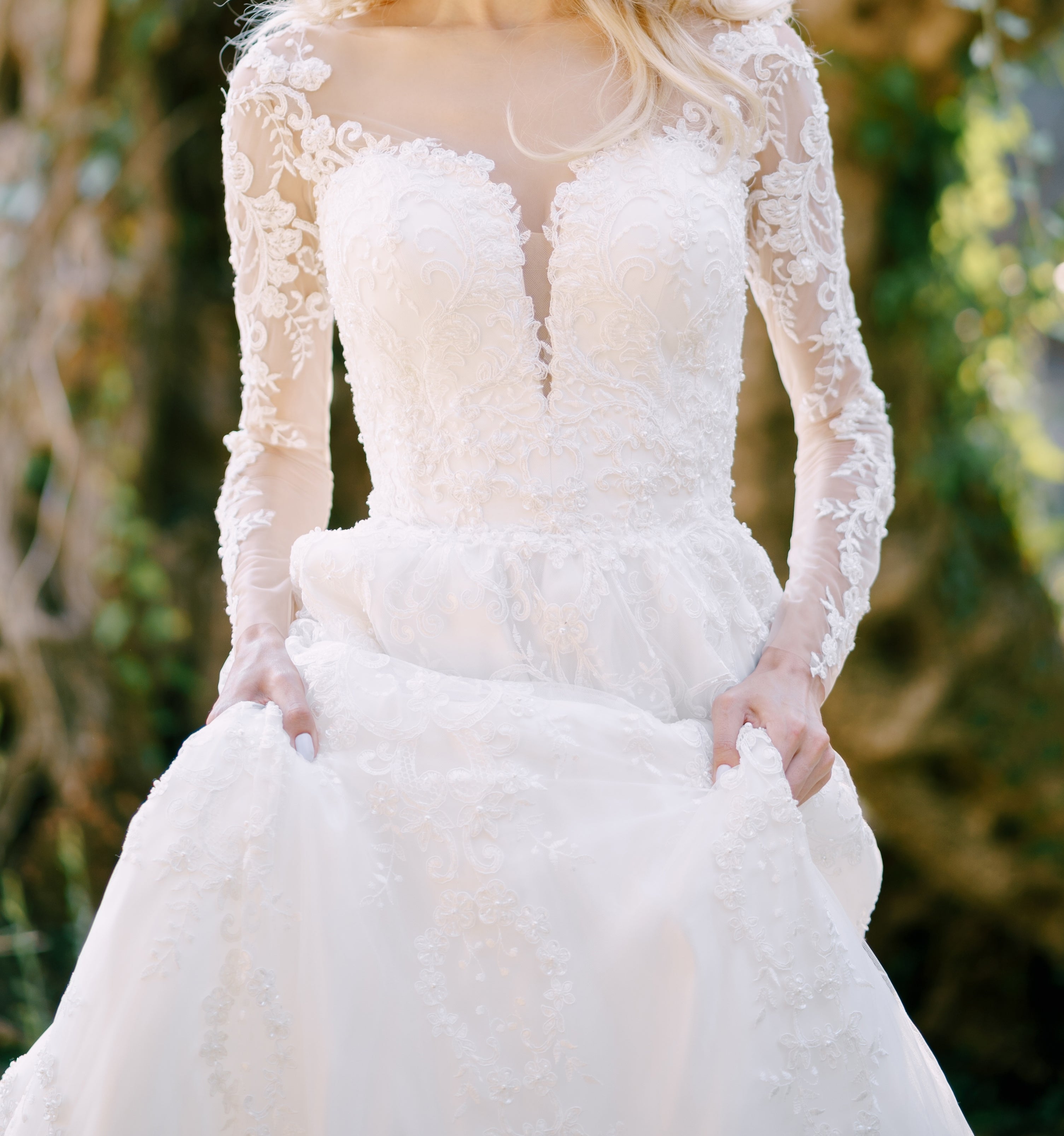 A woman in a lace wedding gown poses outdoors, holding the skirt, with an expression of serene confidence