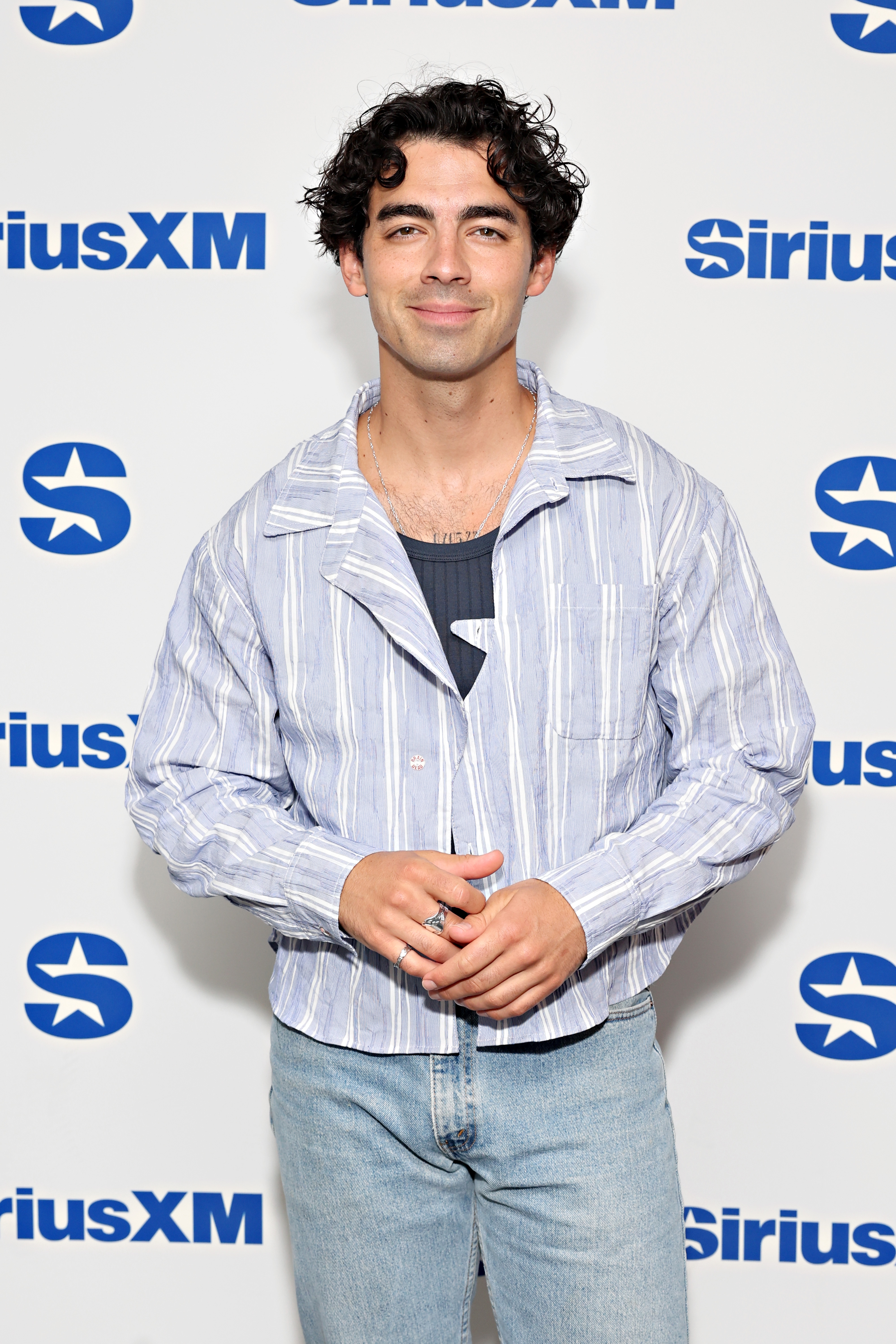 Joe Jonas poses at SiriusXM event in a casual striped shirt and jeans, smiling at the camera