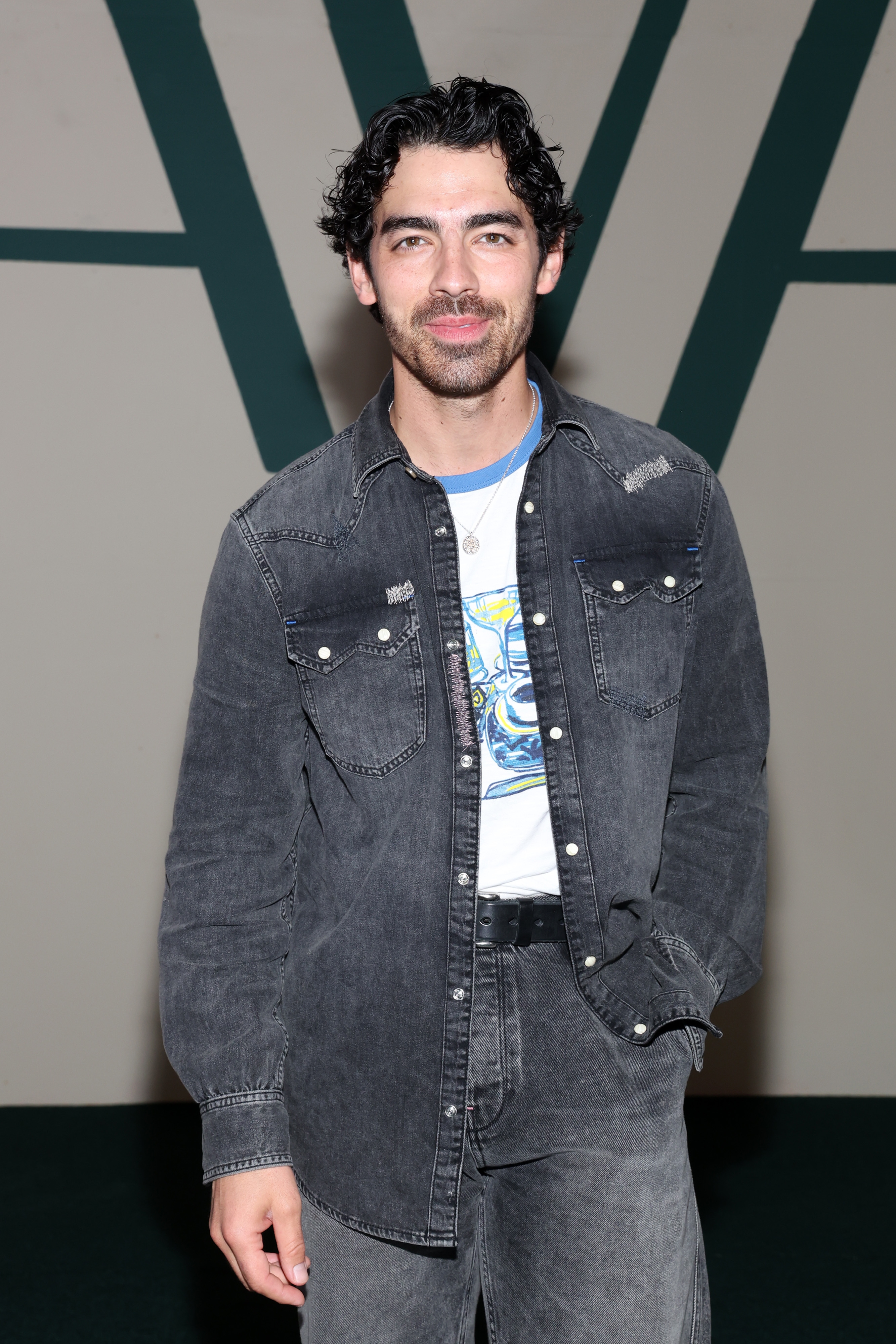Joe Jonas in a denim jacket and jeans stands on a neutral background, smiling at the camera