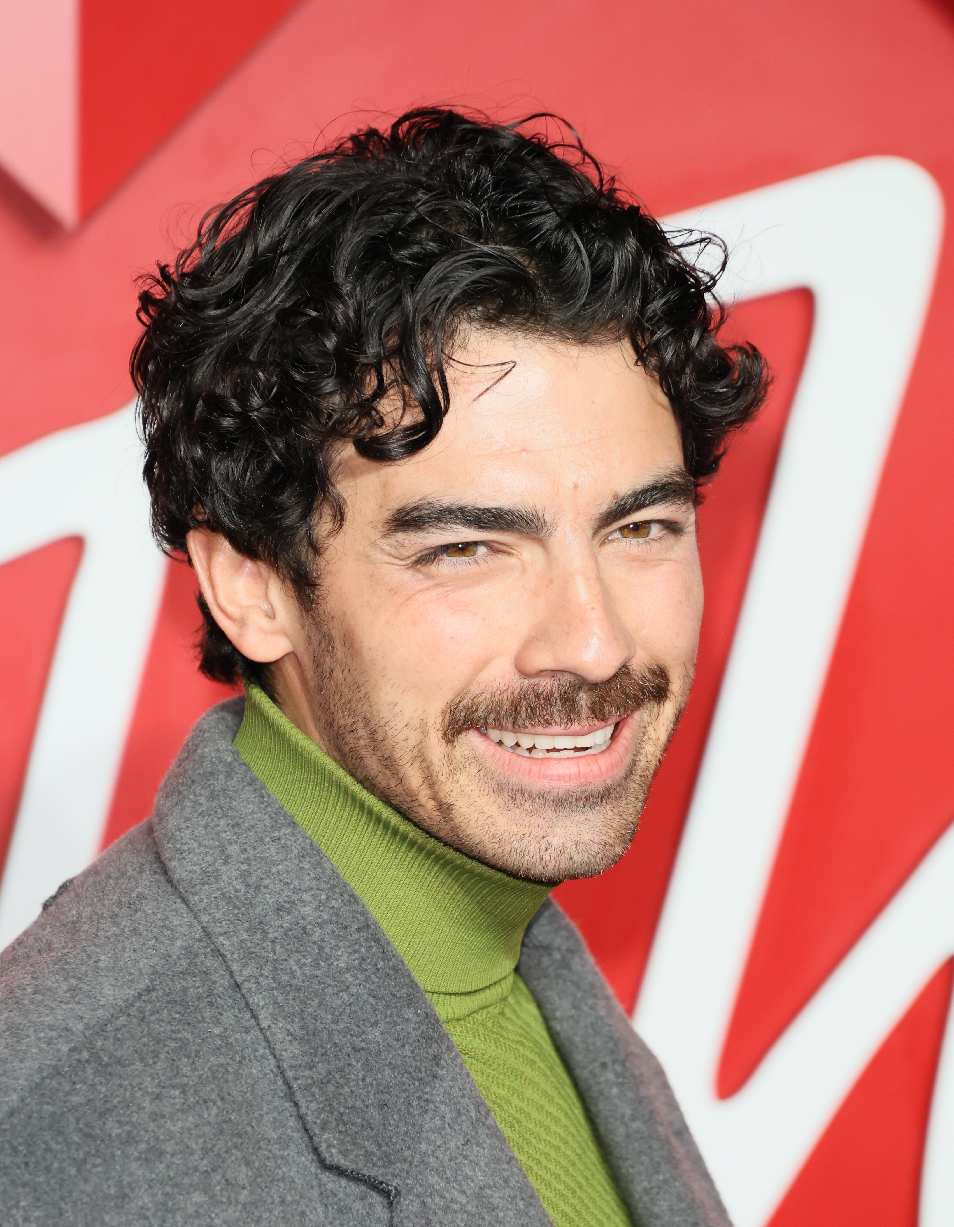 Joe Jonas with curly hair smiles, wearing a gray blazer over a green turtleneck, standing in front of a stylized backdrop