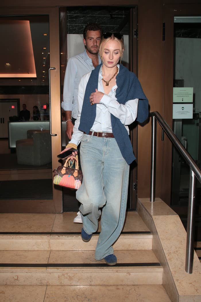 Sophie Turner and Peregrine Pearson in casual outfit with sunglasses on head, carrying a colorful bag, walking down stairs. Another person follows behind