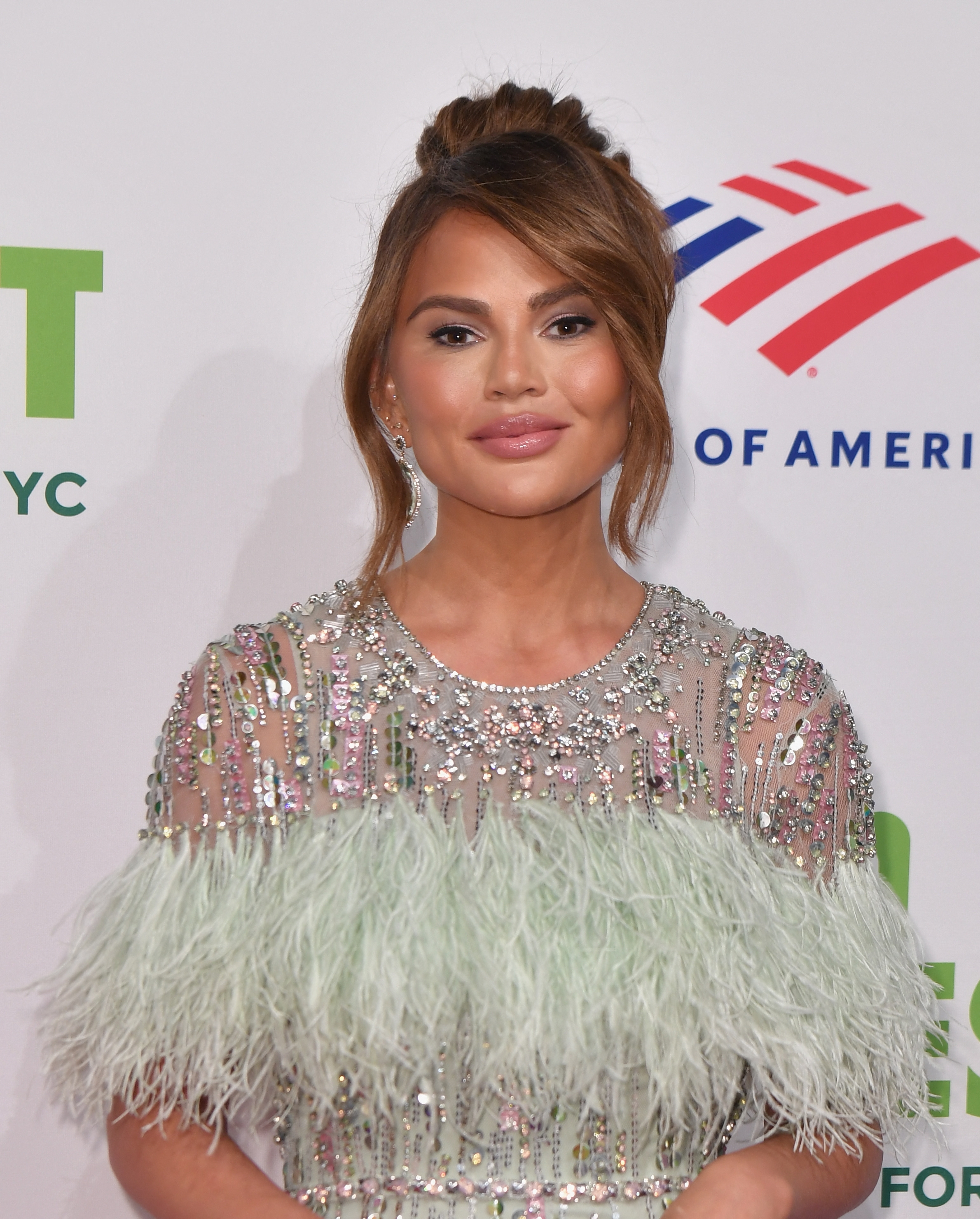 Chrissy Teigen in a feathered, sequined gown on a red carpet, smiling and posing for photos
