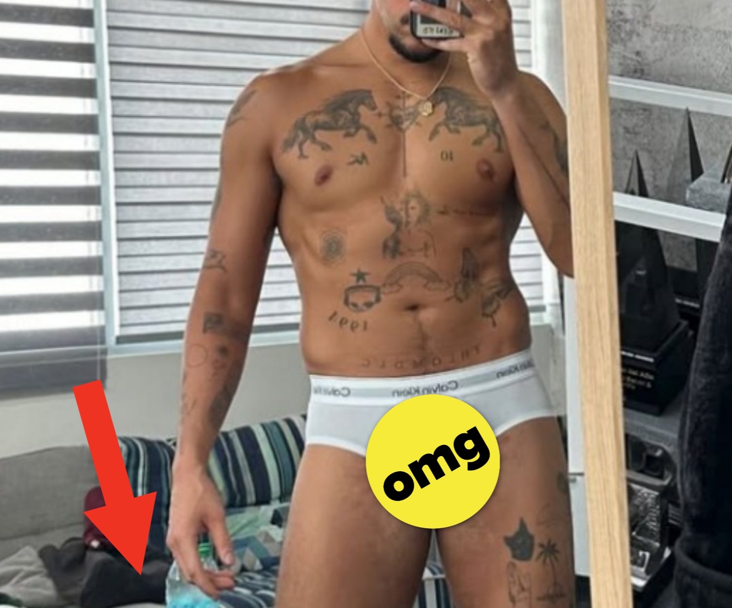 Person takes a mirror selfie wearing underwear, showcasing tattoos. Room has casual clothes and hats in the background