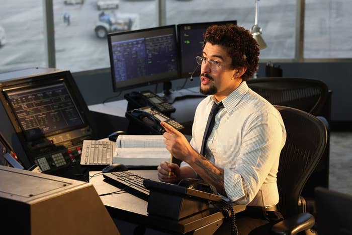 A man in an office speaks on a headset, surrounded by computer monitors and aviation equipment, likely in an air traffic control setting