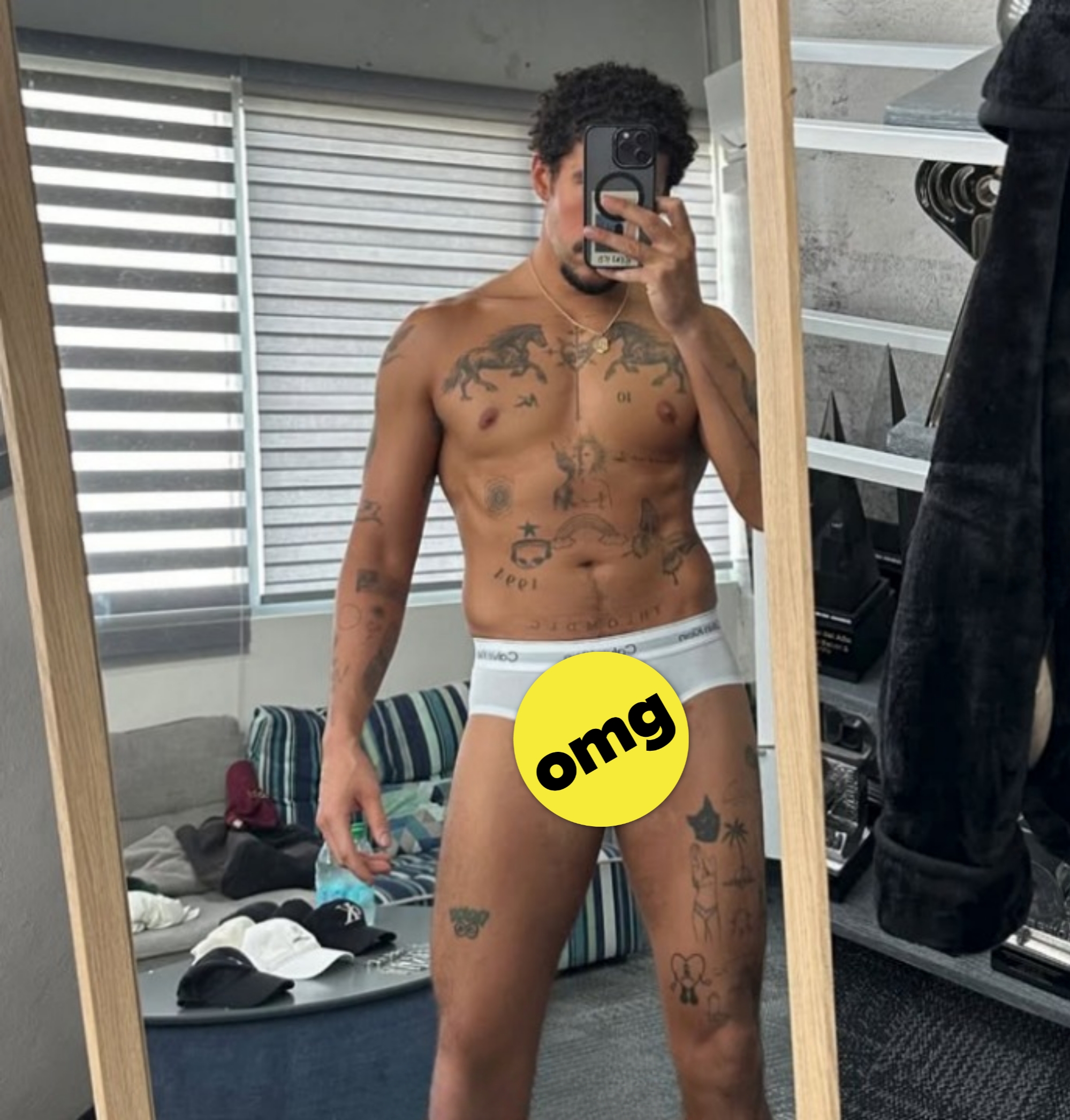Person taking a mirror selfie, showing tattoos and wearing white briefs in a casual home environment