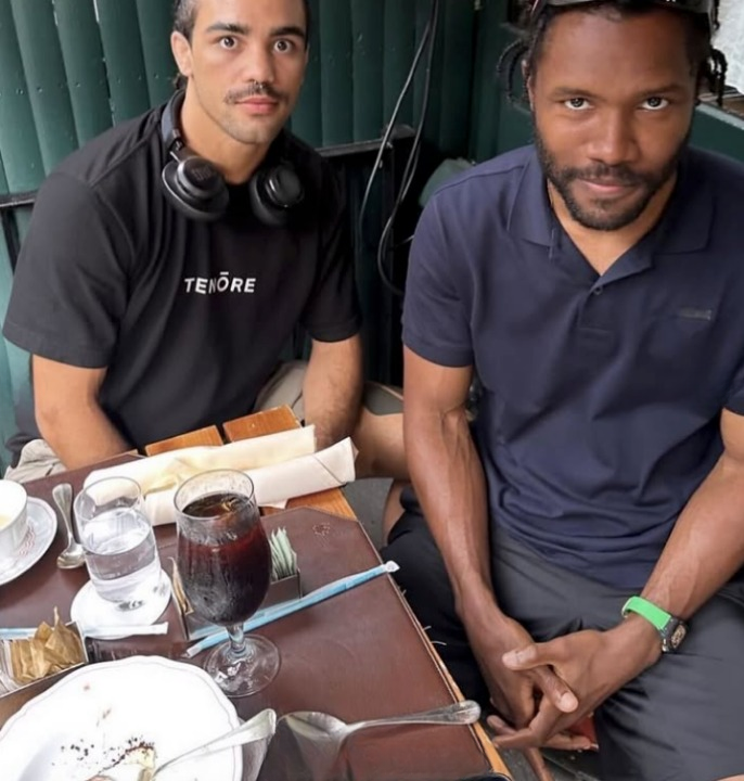 Two people sitting at a table with drinks and plates, one wearing headphones around their neck and a black shirt, the other in a collared shirt