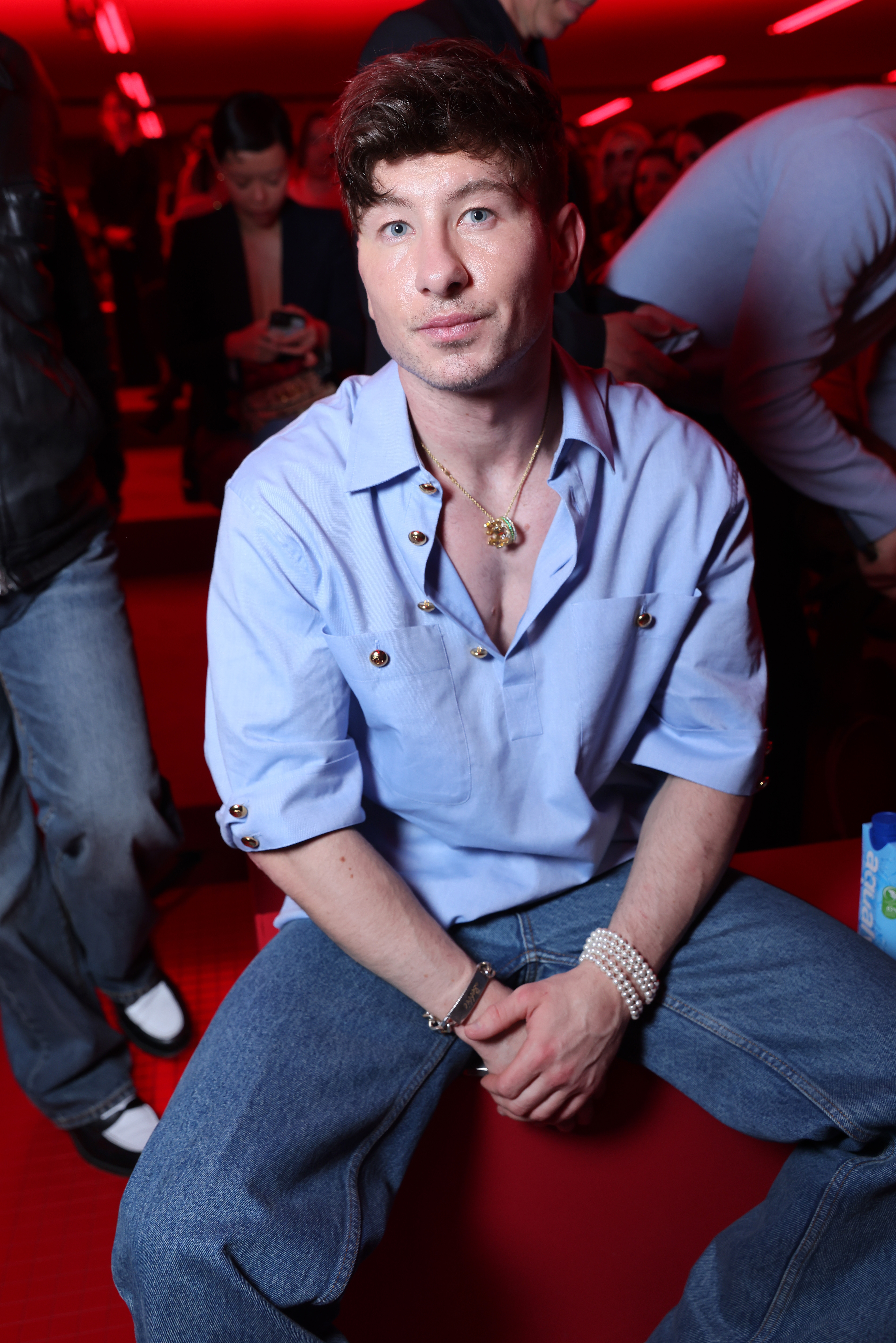 Barry Keoghan seated in a casual event, wearing a light shirt and jeans, with accessories including a necklace and bracelet