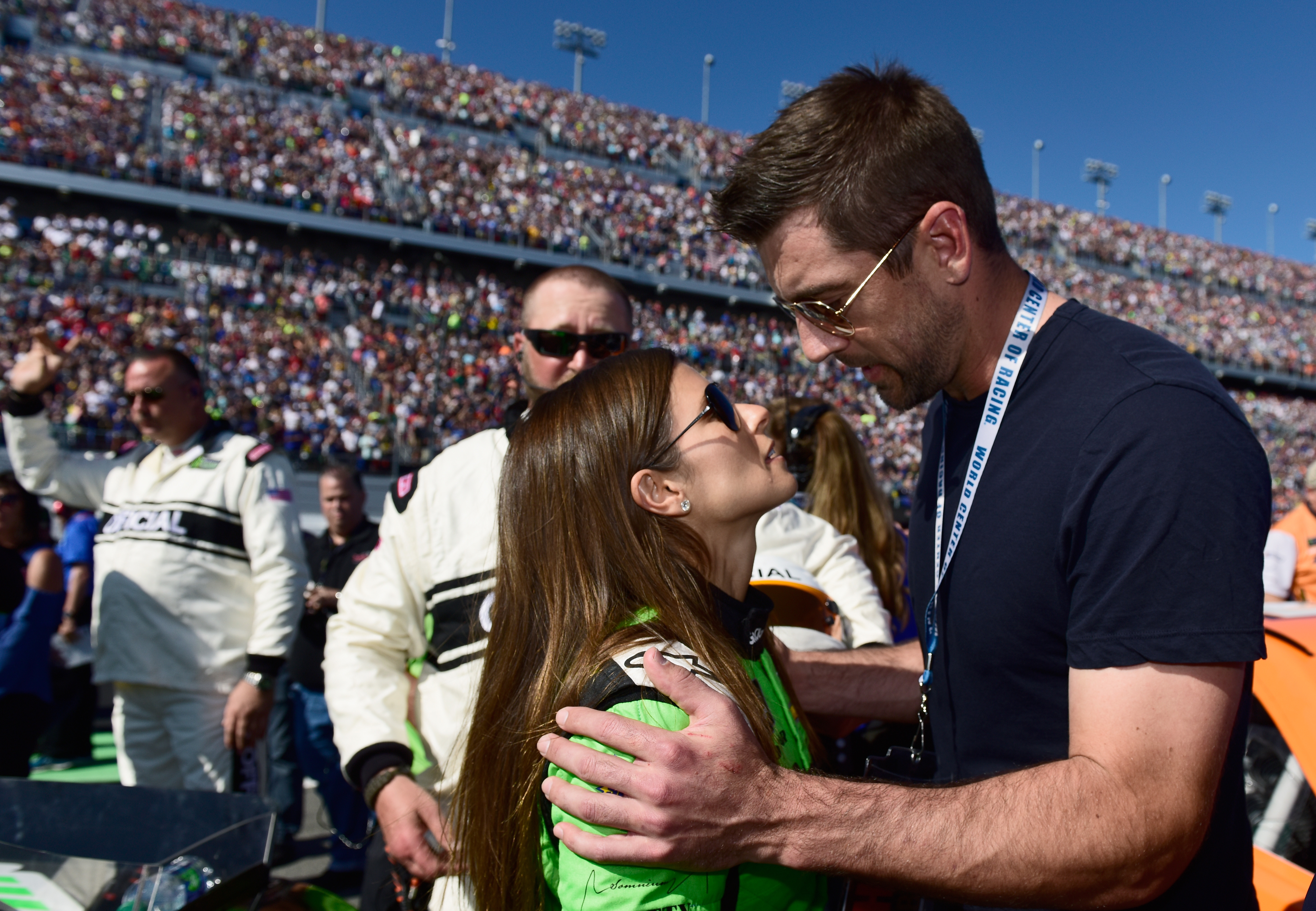 Danica Patrick and Aaron Rodgers in a racing suit share a close moment at a racetrack event with a crowd and other people in the background